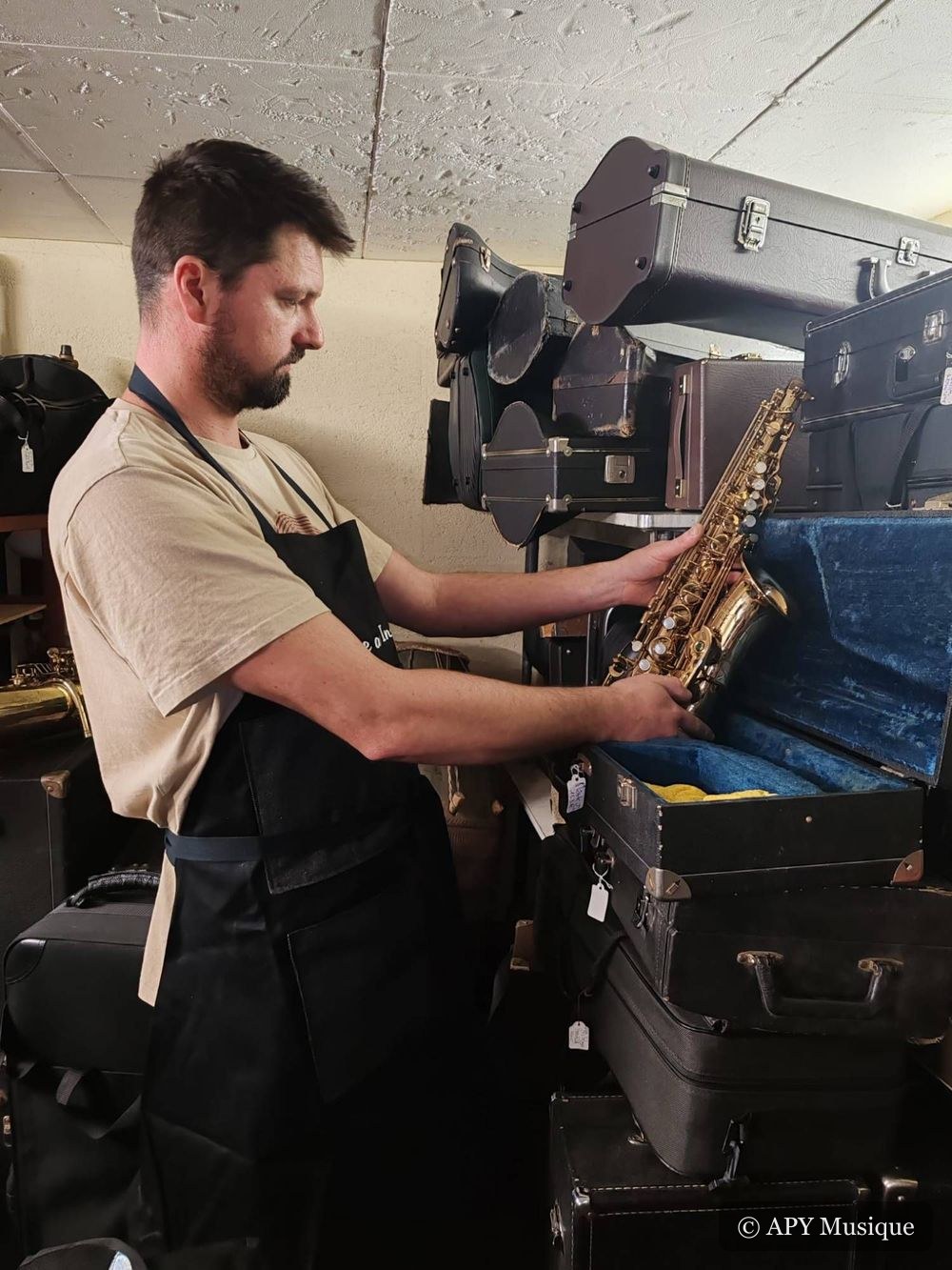 Amaury Plecety sortant un saxophone alto verni de son étui parmi les étuis empilés du stock de l’atelier