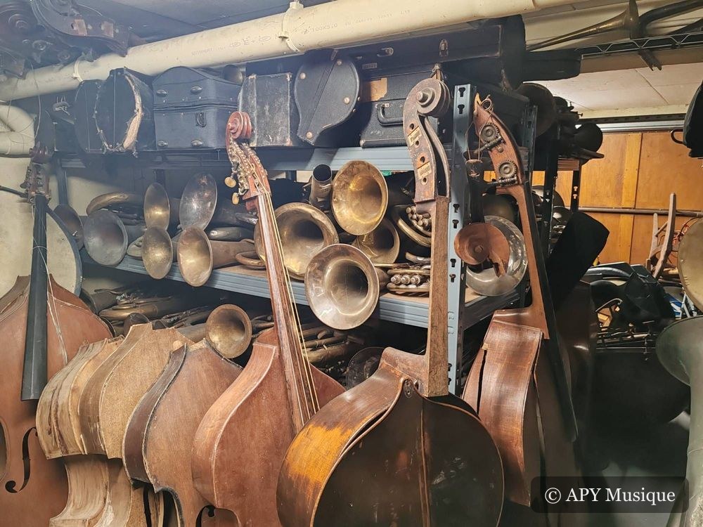 Contrebasses et pavillons de cuivres rangés sur des rayonnages dans la réserve de l’atelier